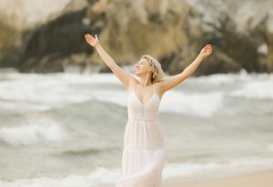 happy woman dances with arms raised spinning dancing on the beach at the ocean wearing a white dress large brown rocks in background