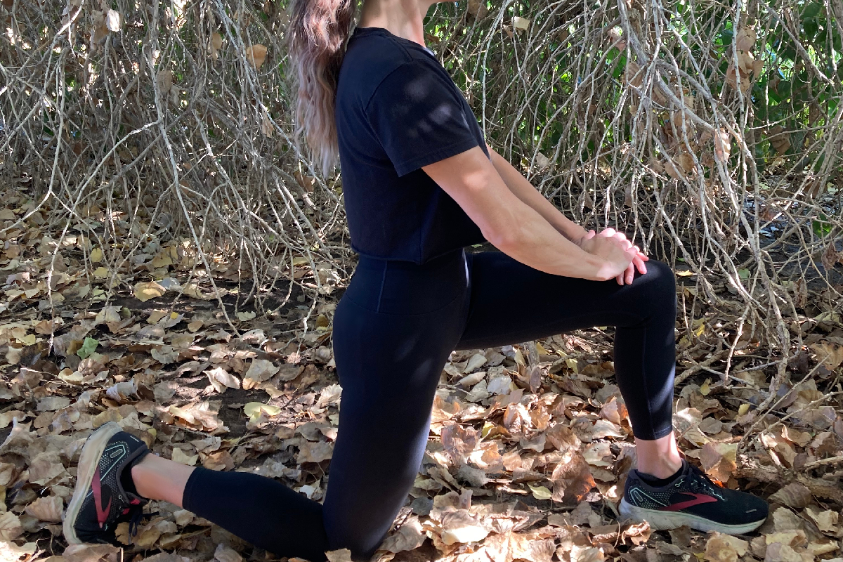 woman kneeling like she is proposing with hands clasped and fingers interlocked resting on top of front knee while back leg is down at a ninety degree angle behind her black beautiful outfit brown leaves contrast around her