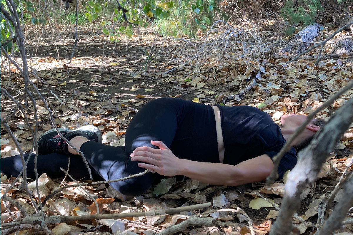 girl stretching torso knee all the way down to the ground rotation black pants and tshirt blond hair black workout shoes leaves fall summer plants tree branches healthy alive
