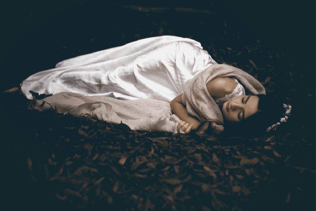 beautiful brunette woman sleeping on her side facing camera eyes closed peacefully on forest floor of dried autumn brown leaves wearing a white robe wrapped in off white shawl with a white pearl tiara or headband in her hair